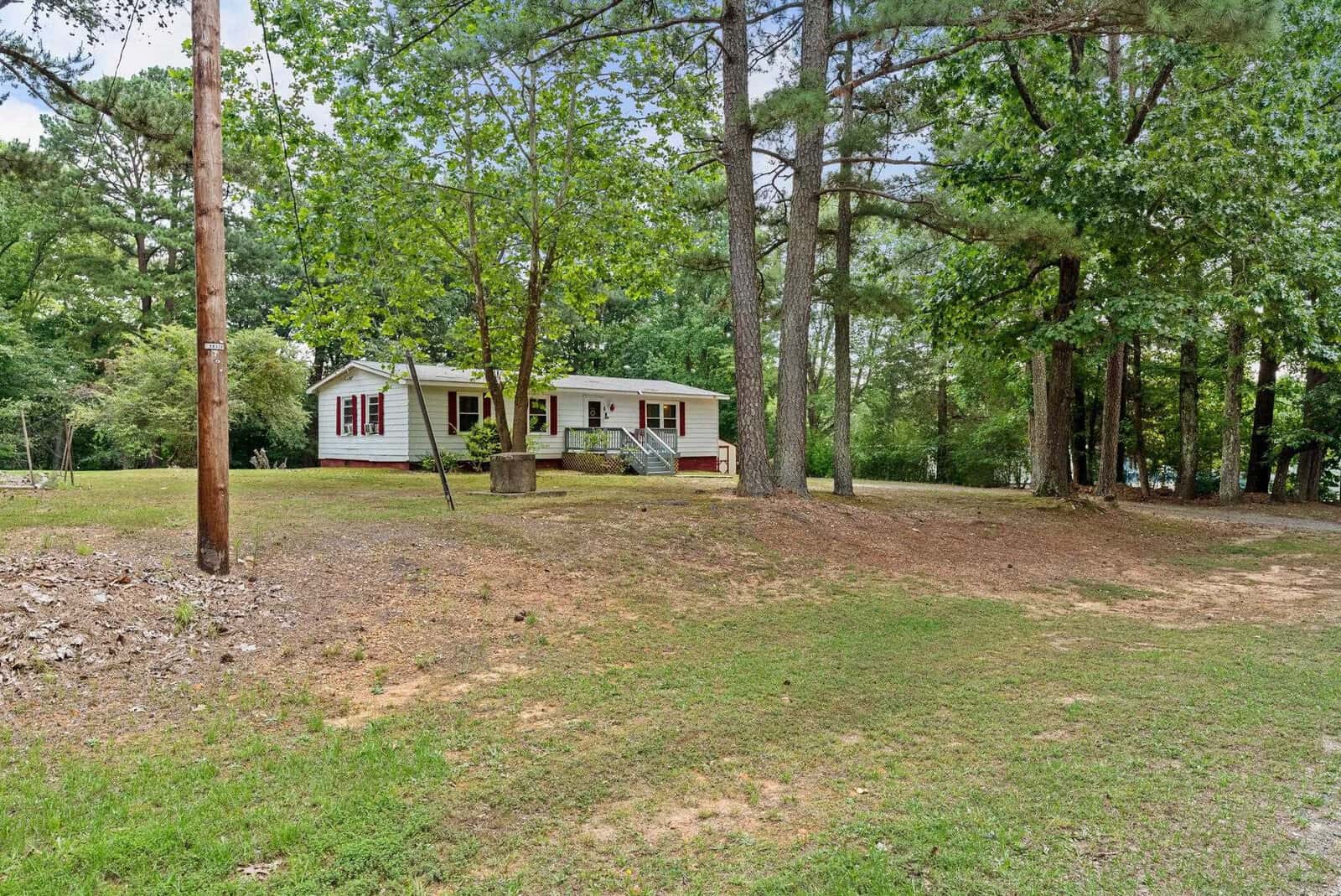 A single-story house with white siding and red trim nestled among tall green trees in a spacious yard with a partly grassy and partly bare ground.