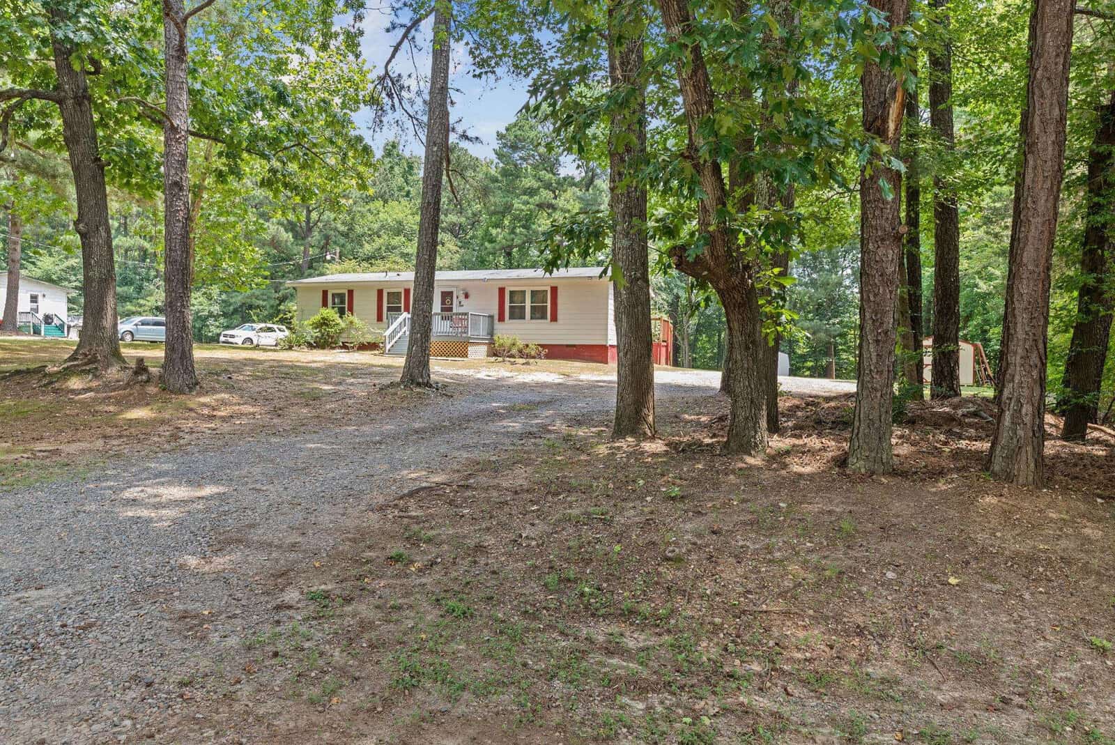 A single-story house with a beige exterior and white trim, surrounded by a wooded area with several trees and a gravel driveway.