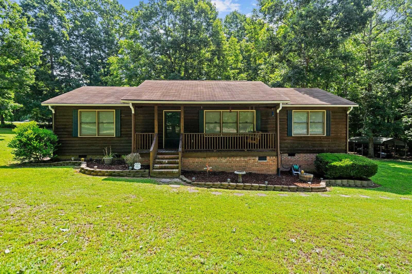A single-story brown house with a shingled roof, located at 1355 Rock Mill Road, Henderson NC, featuring a front porch with railings and steps, surrounded by a well-maint