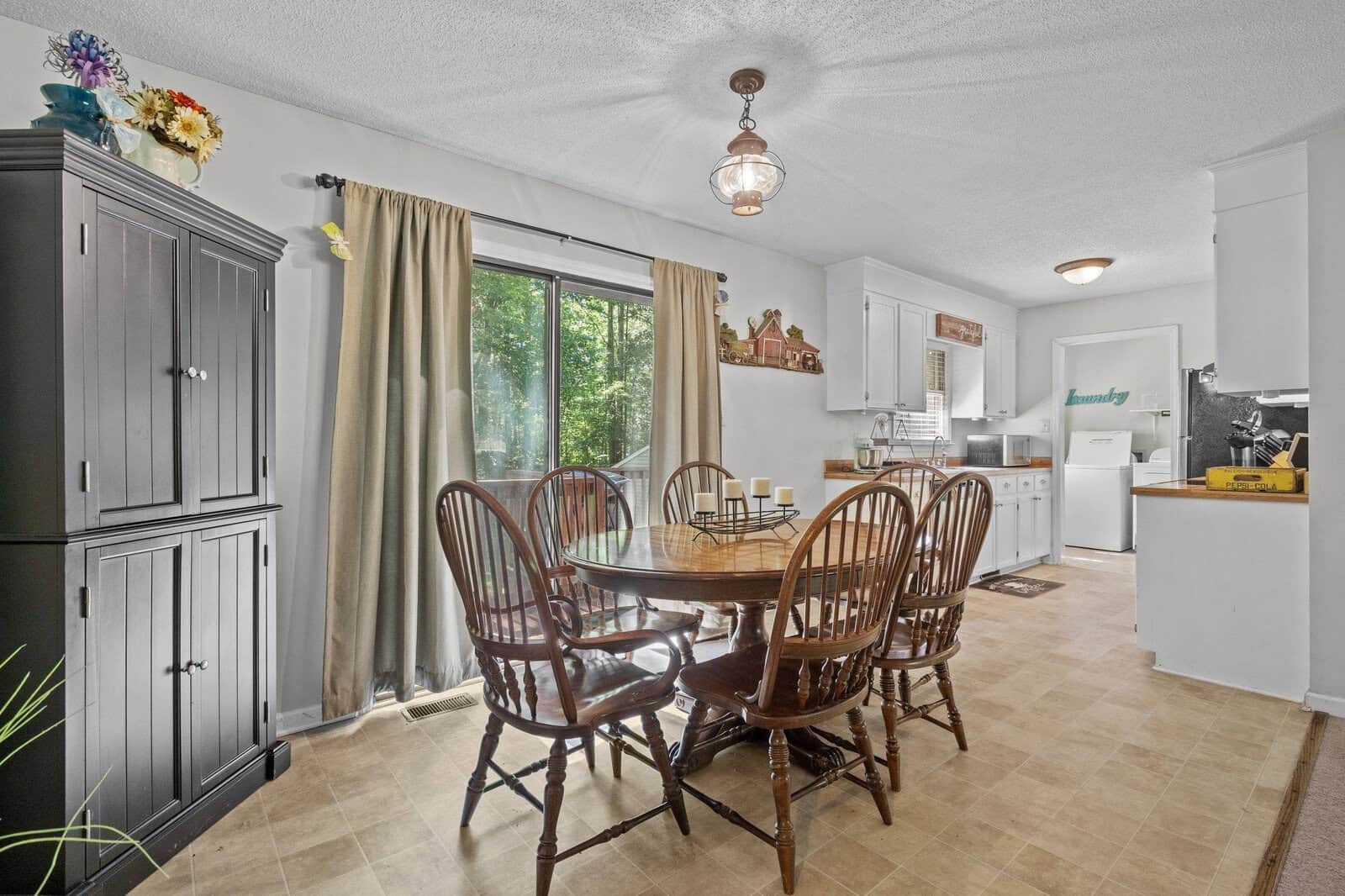 A well-lit dining area with a wooden round table and chairs, adjacent to a kitchen with modern appliances and cabinets.