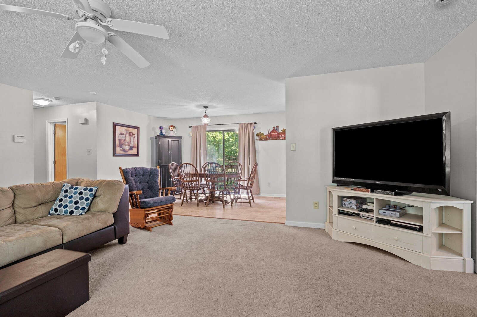 A well-lit, carpeted living room featuring a large tv stand, a sectional sofa, and a dining area with a wooden table set visible in the background.