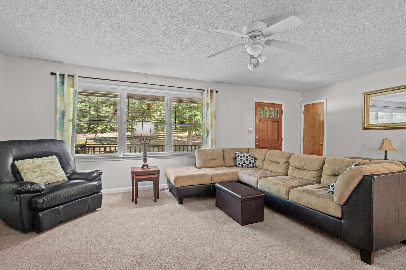 A well-lit living room featuring a sectional beige sofa with patterned cushions, a black leather armchair, an ottoman, and a ceiling fan above. the room has a large window with green curtains, a lamp on a small table, and a carpeted floor. a wooden front door with glass panels is visible in the background.