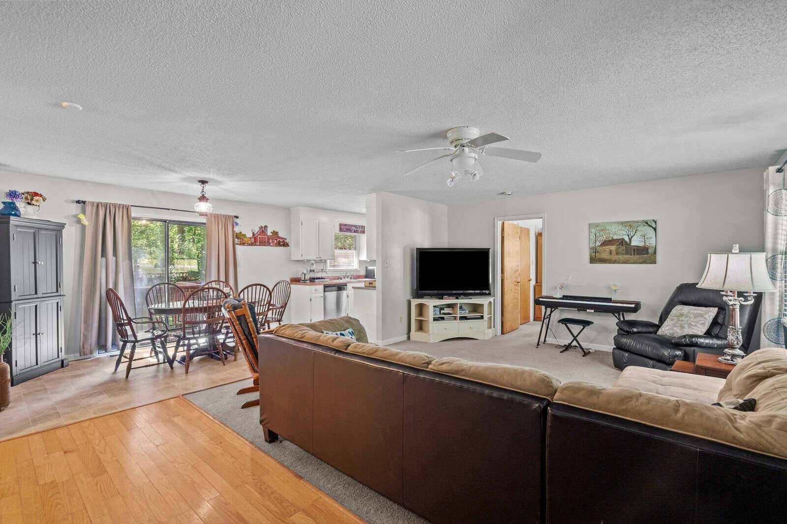 A well-lit, open-plan living space featuring a hardwood floor, with a seating area that includes a brown sofa and armchair, a television on a stand, and a ceiling fan above. to the side is a dining area with a wooden table and chairs, adjacent to a kitchen with white cabinets. the room is accessorized with various home decor items, and natural light streams in through the sliding glass doors leading to an outdoor space.