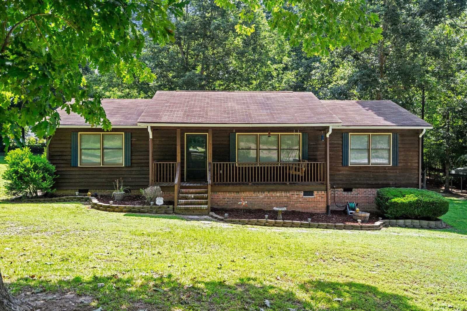 A single-story house with brown siding, a red shingled roof, and a front porch, surrounded by green lawn and trees under a clear sky.