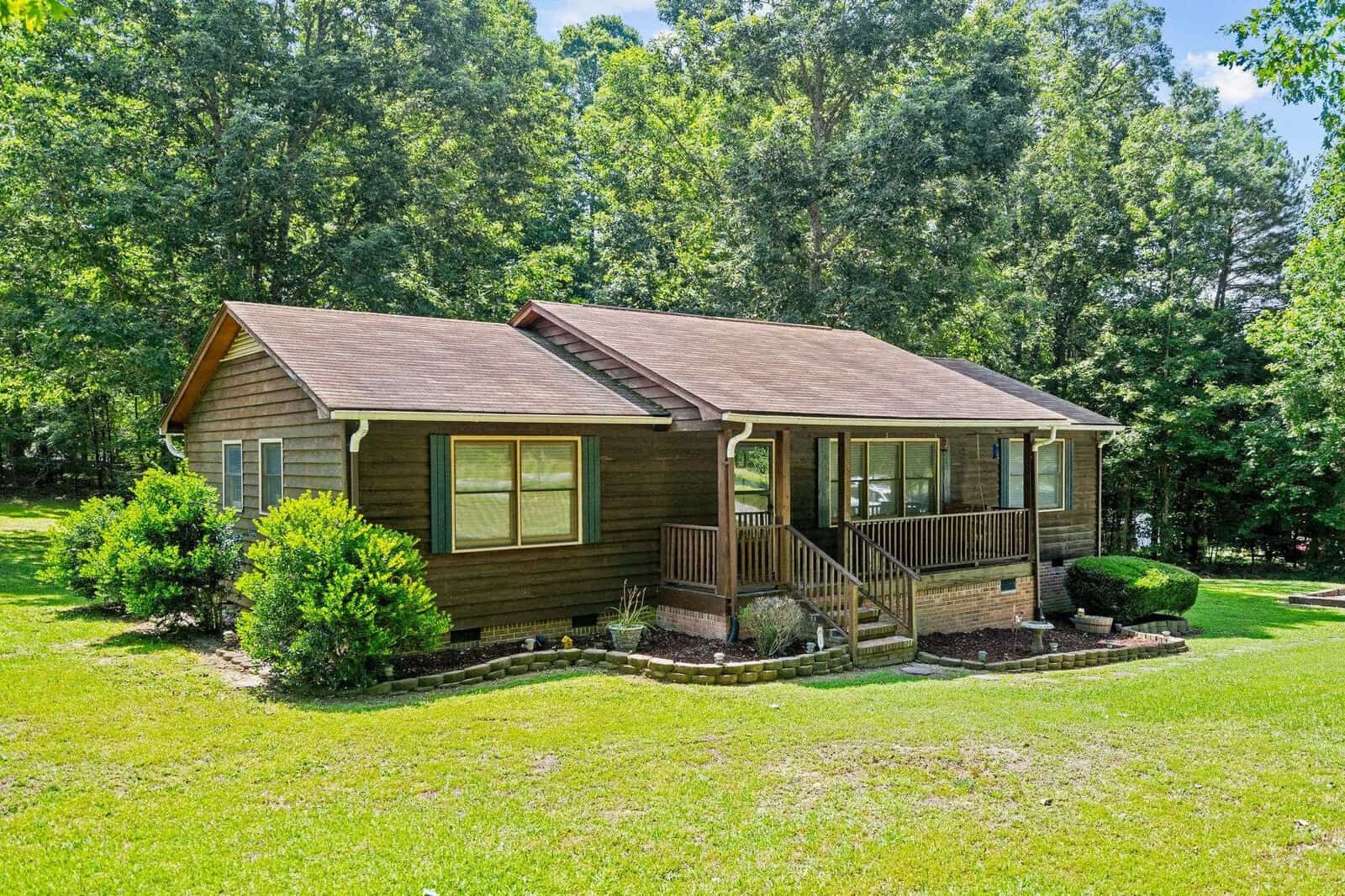 A single-story brown house with a gable roof, featuring large front windows and a small porch, set against a backdrop of green trees under a clear sky.