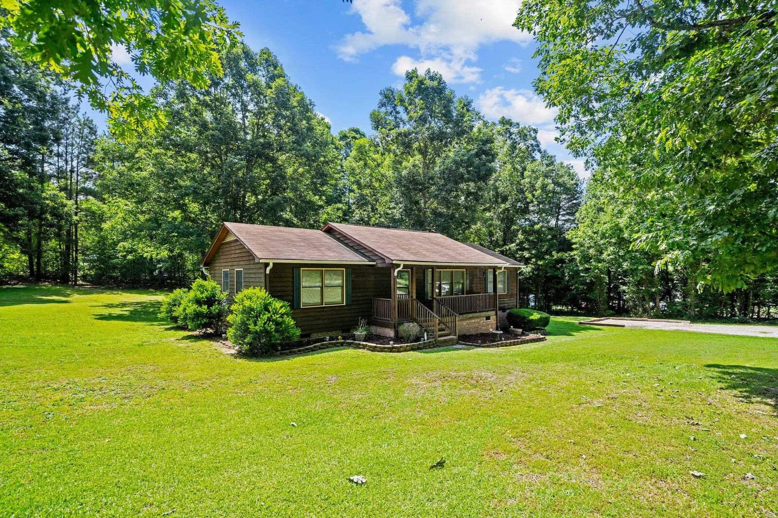 A single-story house with a brown wooden exterior, surrounded by a lush lawn and dense green trees under a clear sky.