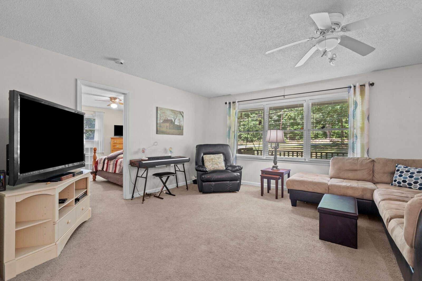 A spacious living room with beige carpeting, featuring a large sectional sofa, a black recliner, a flat-screen television on a white stand, an ironing board, and a ceiling fan above, with natural light streaming in through windows covered with patterned curtains.