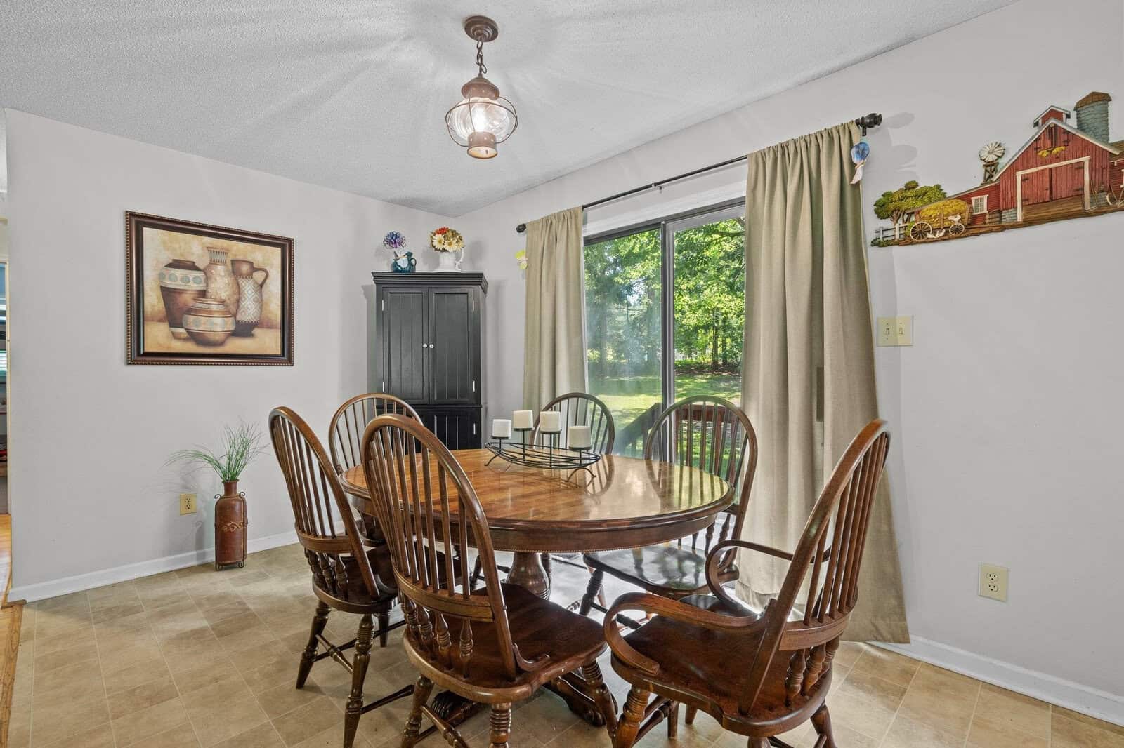 A well-lit dining room featuring a wooden table with chairs, a black cabinet, and decorative items on the walls, with a view of greenery outside the window.