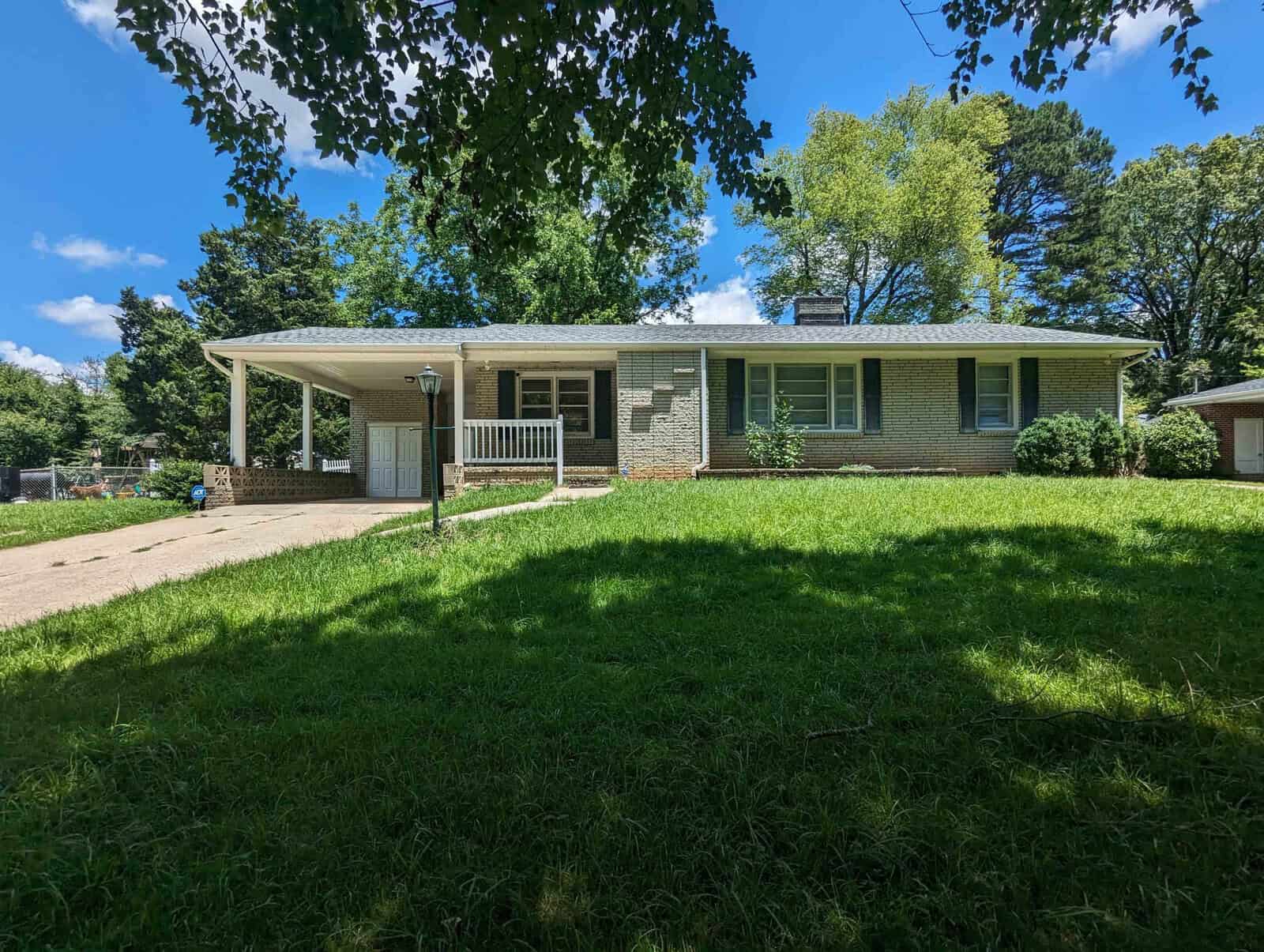 A single-story house with a brick façade, a carport on the left, and a small porch, surrounded by a lawn with trees in the background on a sunny day.