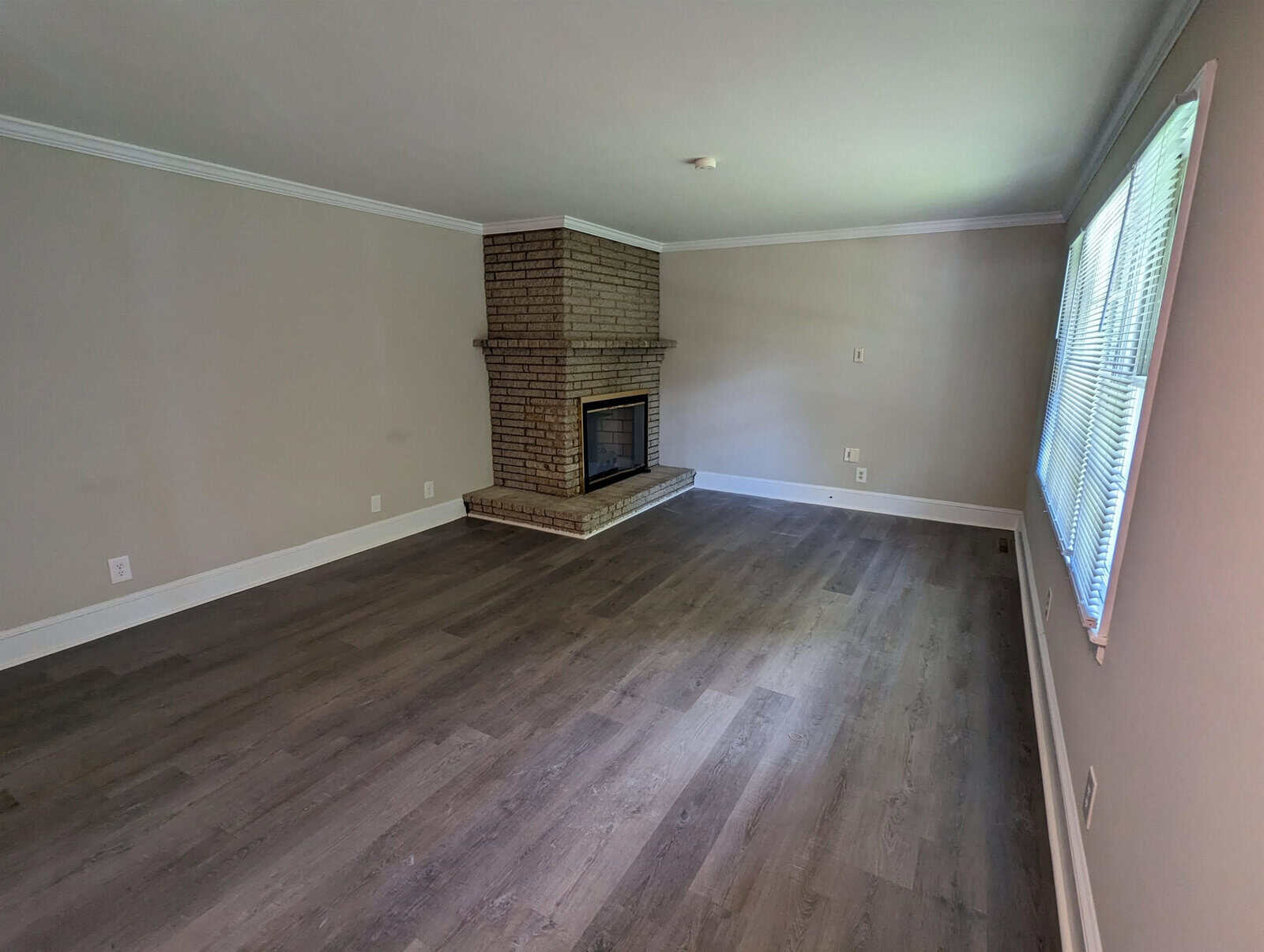 An empty room featuring a brick fireplace on the right, with laminate flooring, white walls, and a window with blinds on the left side.