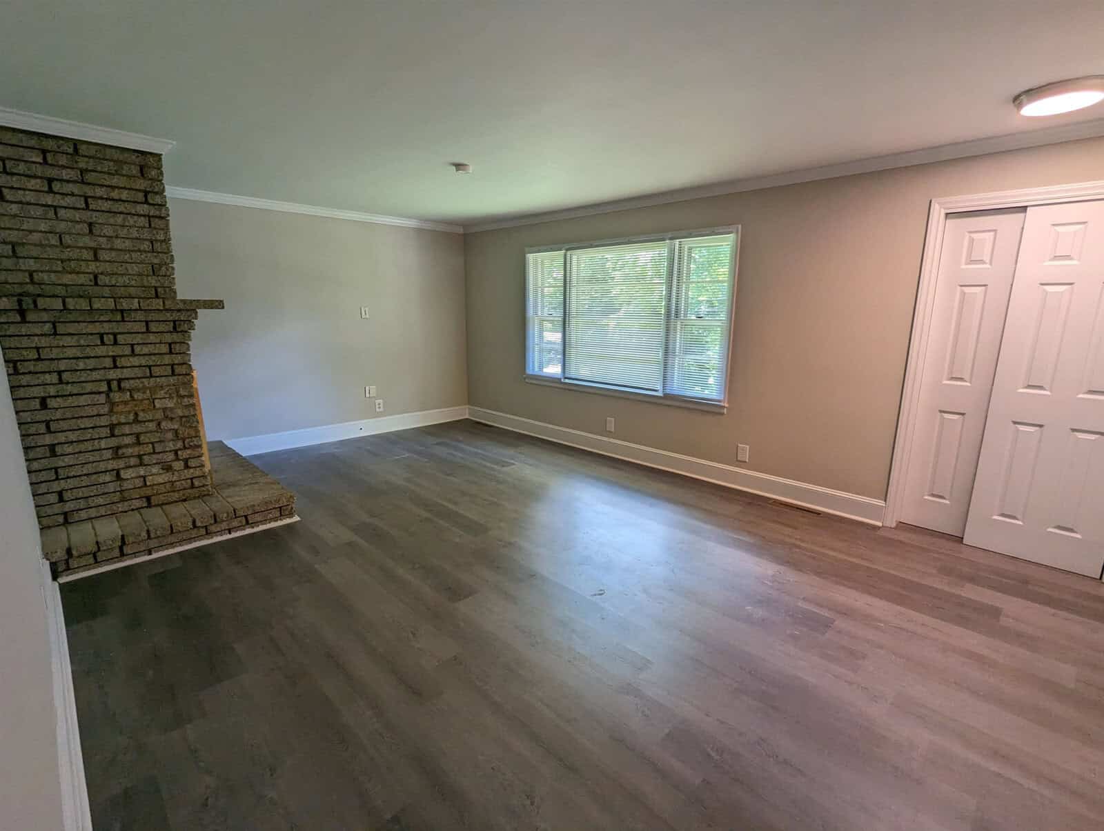 A spacious, empty room with laminate flooring, featuring a brick fireplace on the left and a large window with a view of greenery. the room has neutral-colored walls and white trim, with a closed door on the right side.