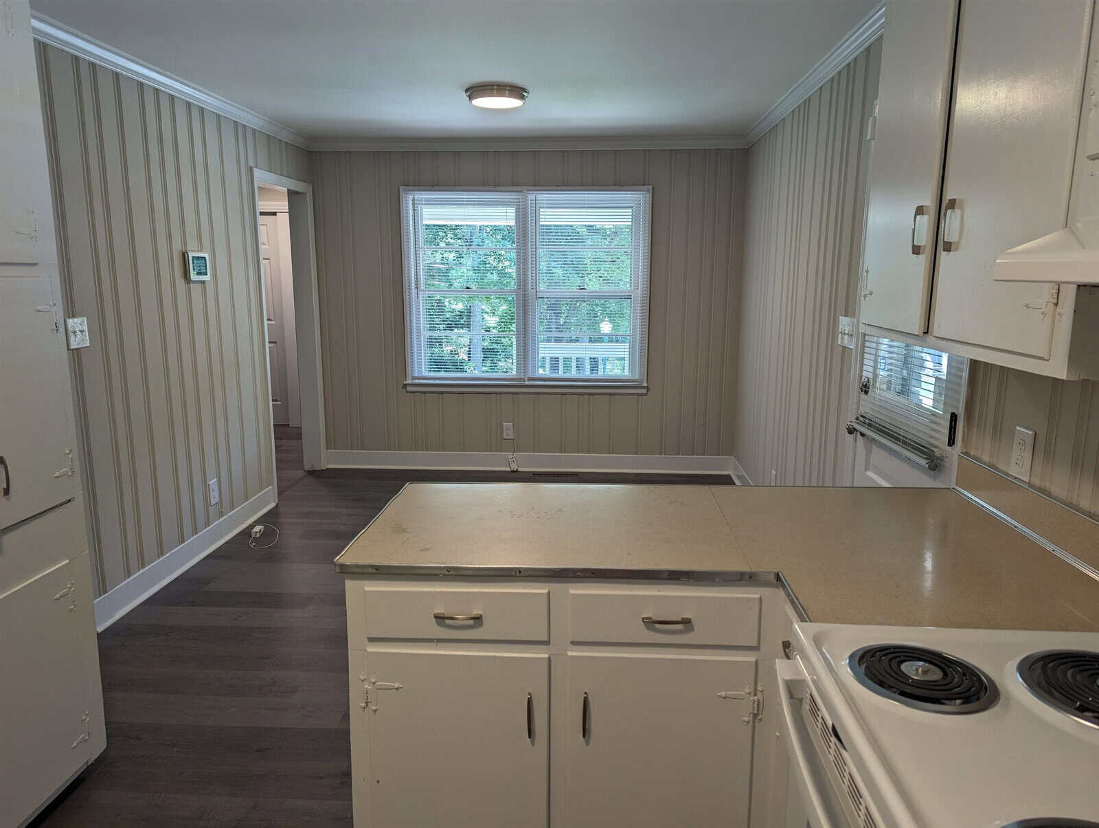 A small kitchen with white cabinetry, beige countertops, a white stove, and grey flooring illuminated by natural light from a window.