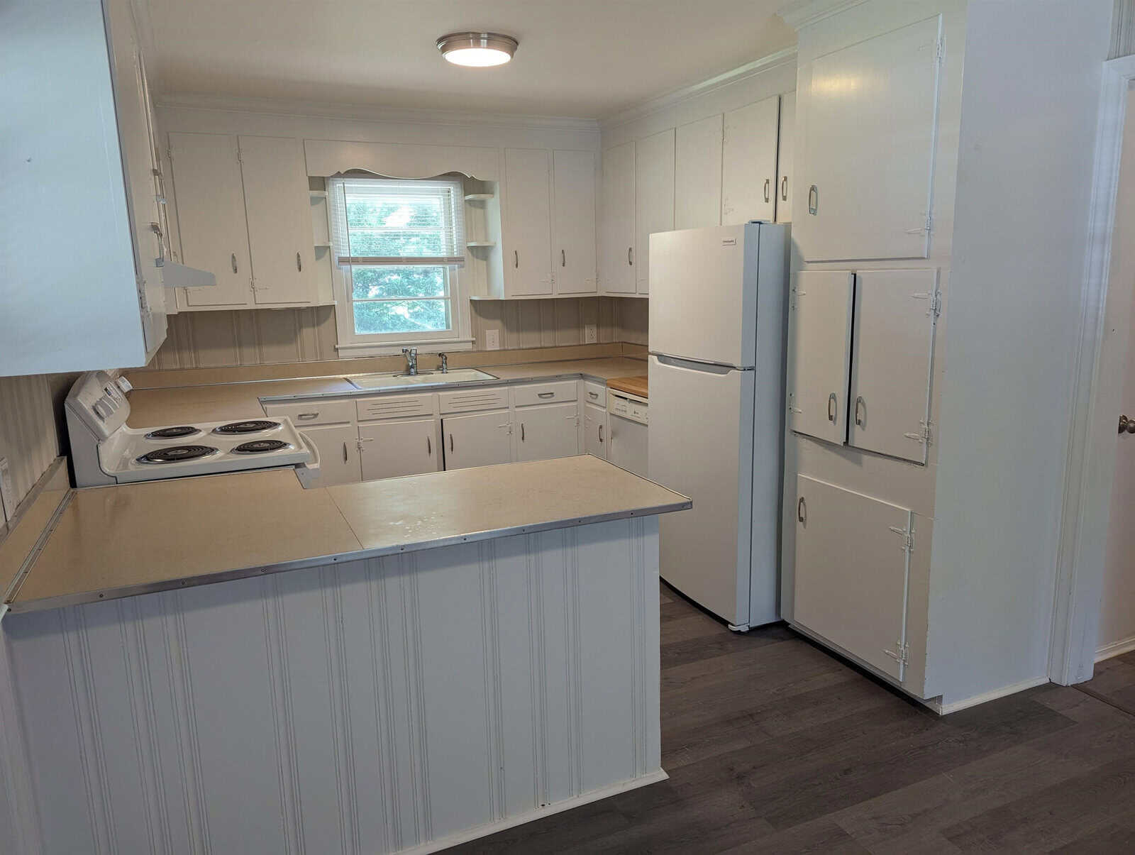 A small, narrow kitchen with white cabinetry, white appliances, and a beige countertop, featuring a window above the sink.