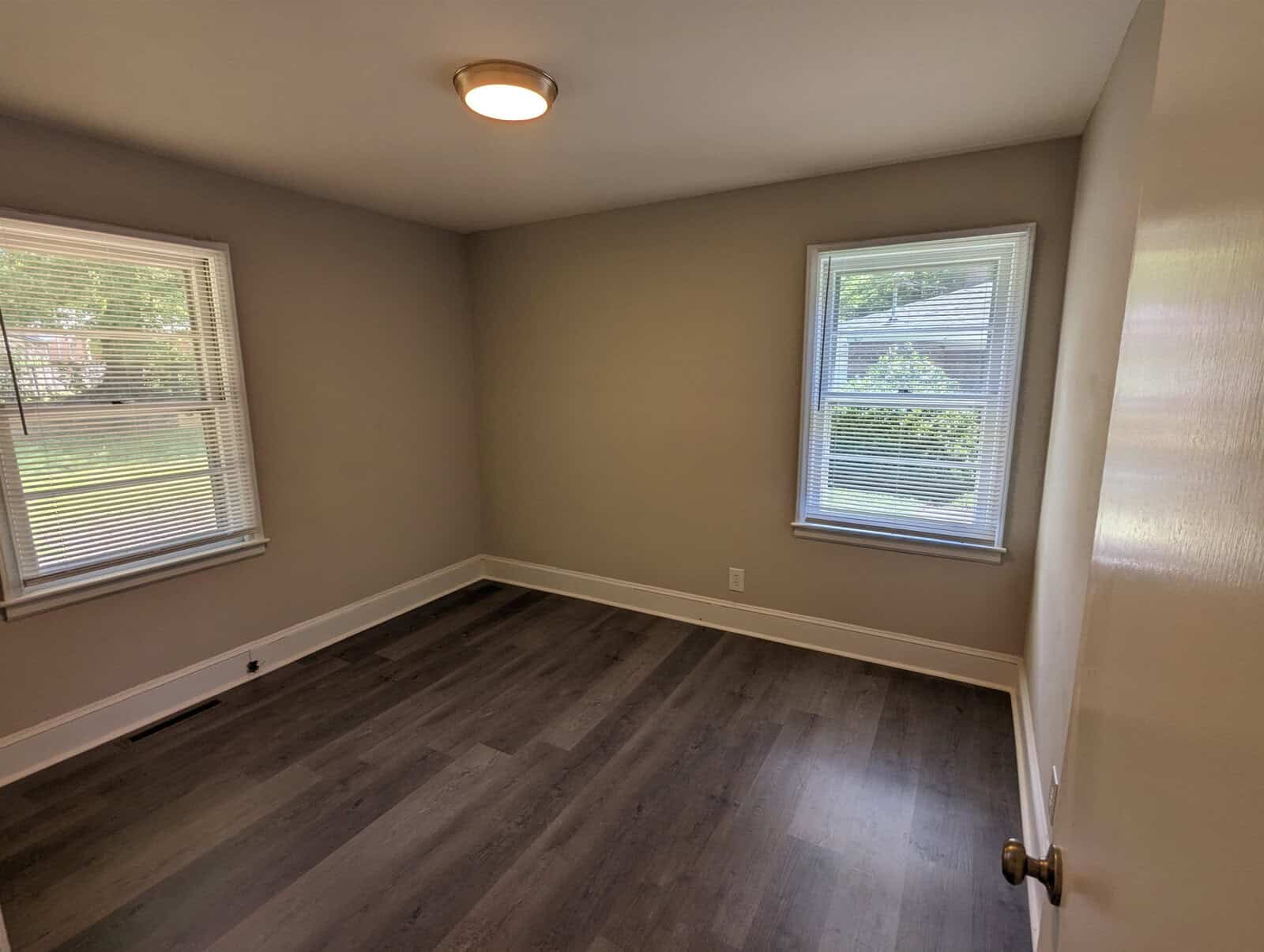 An empty room with grey walls, dark laminate flooring, two windows with blinds, and a single ceiling light fixture.