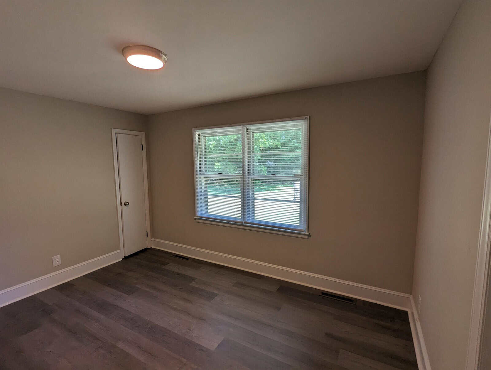 An empty room with gray floors, one door, and a large window with blinds overlooking greenery outside.