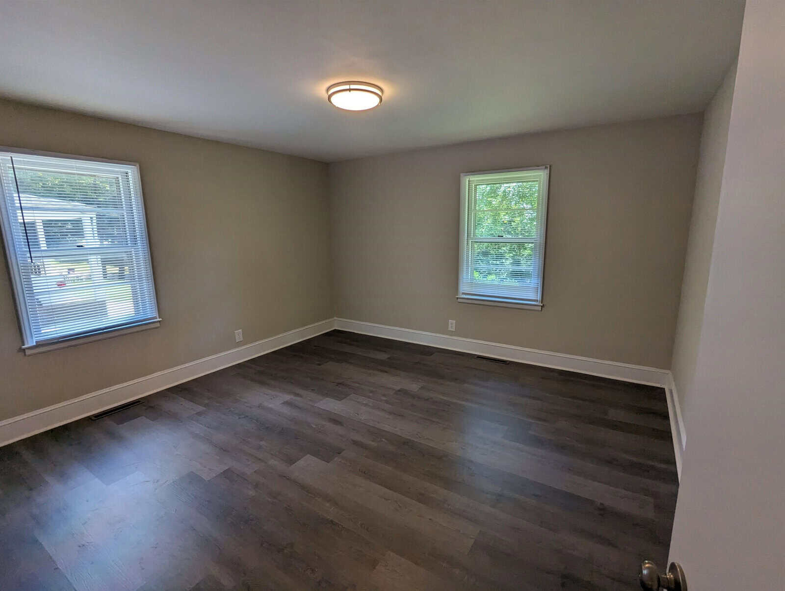 An empty room with gray walls, dark wood laminate flooring, and two windows providing natural light. there is a central ceiling light fixture.