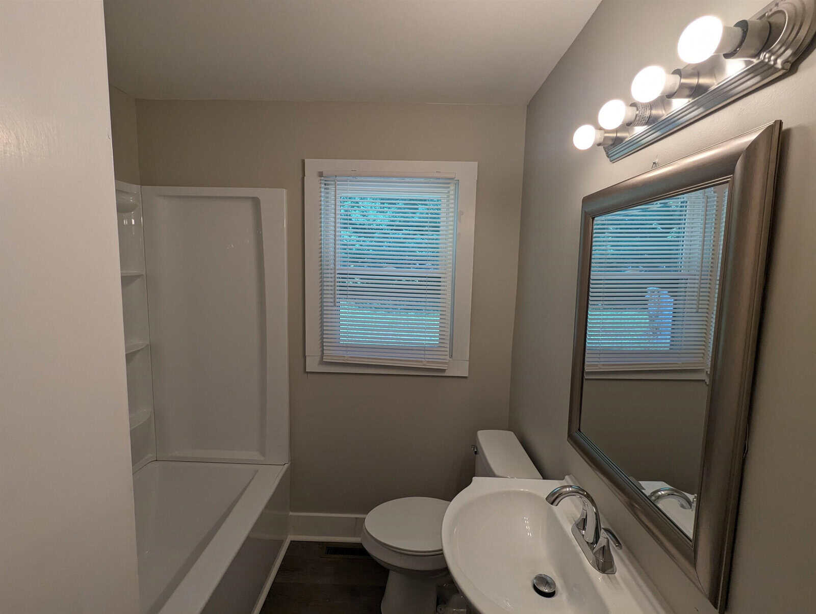 A modern bathroom interior featuring a white bathtub and shower unit, a toilet, a sink with a large mirror and bright overhead lights, and a window with closed blinds.