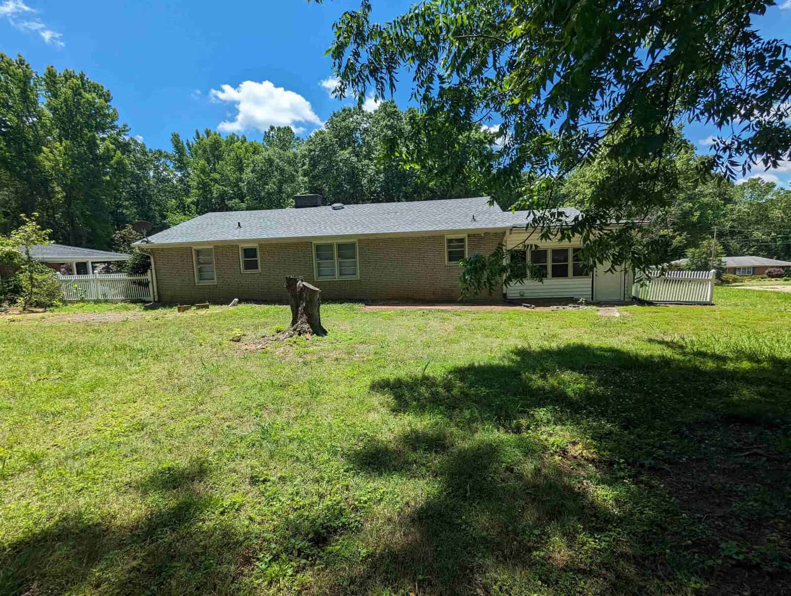 A single-story brick house with a white fence and a tree stump in the front yard on a sunny day.