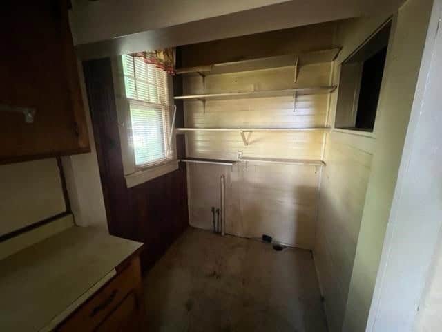 An empty, dimly lit corner of a room with old, unpainted wooden shelving and cabinets, next to a window with a partially closed blind.