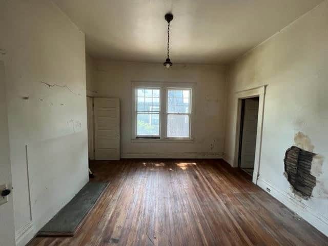 An empty room with wooden floors, peeling paint on the walls, a single window allowing in natural light, and two closed doors.