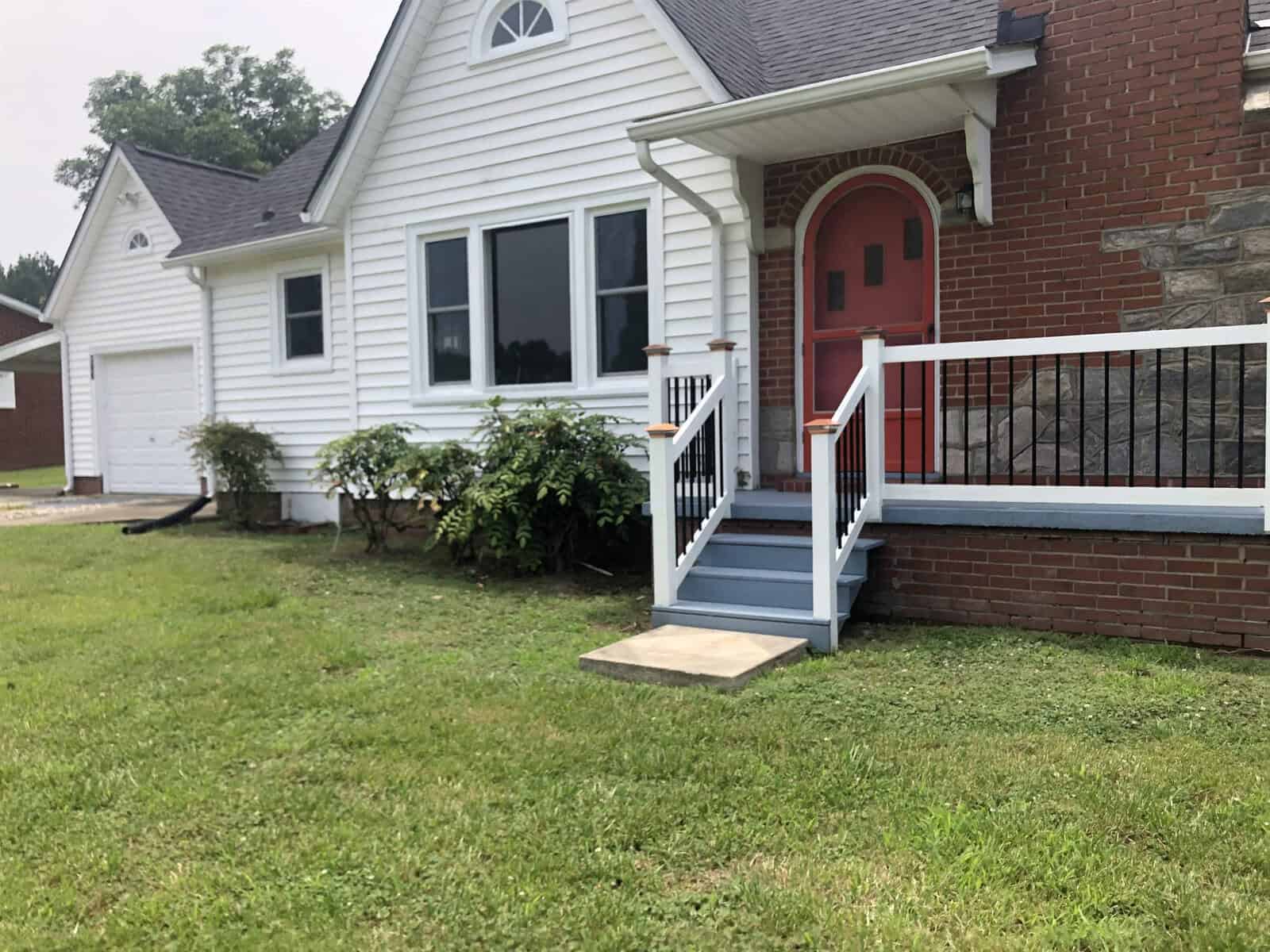 A white single-story home with a red door, flanked by red railings on the porch steps, and a small brick section on its right side, situated on a lawn with visible greenery near the windows.