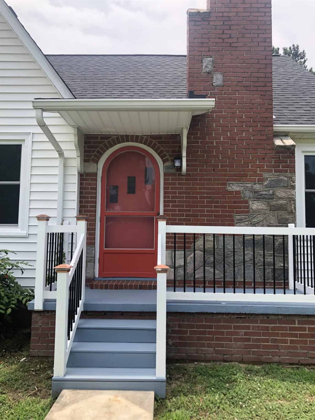 A red front door on a white house with a brick chimney, flanked by a small porch with gray steps and white railings.