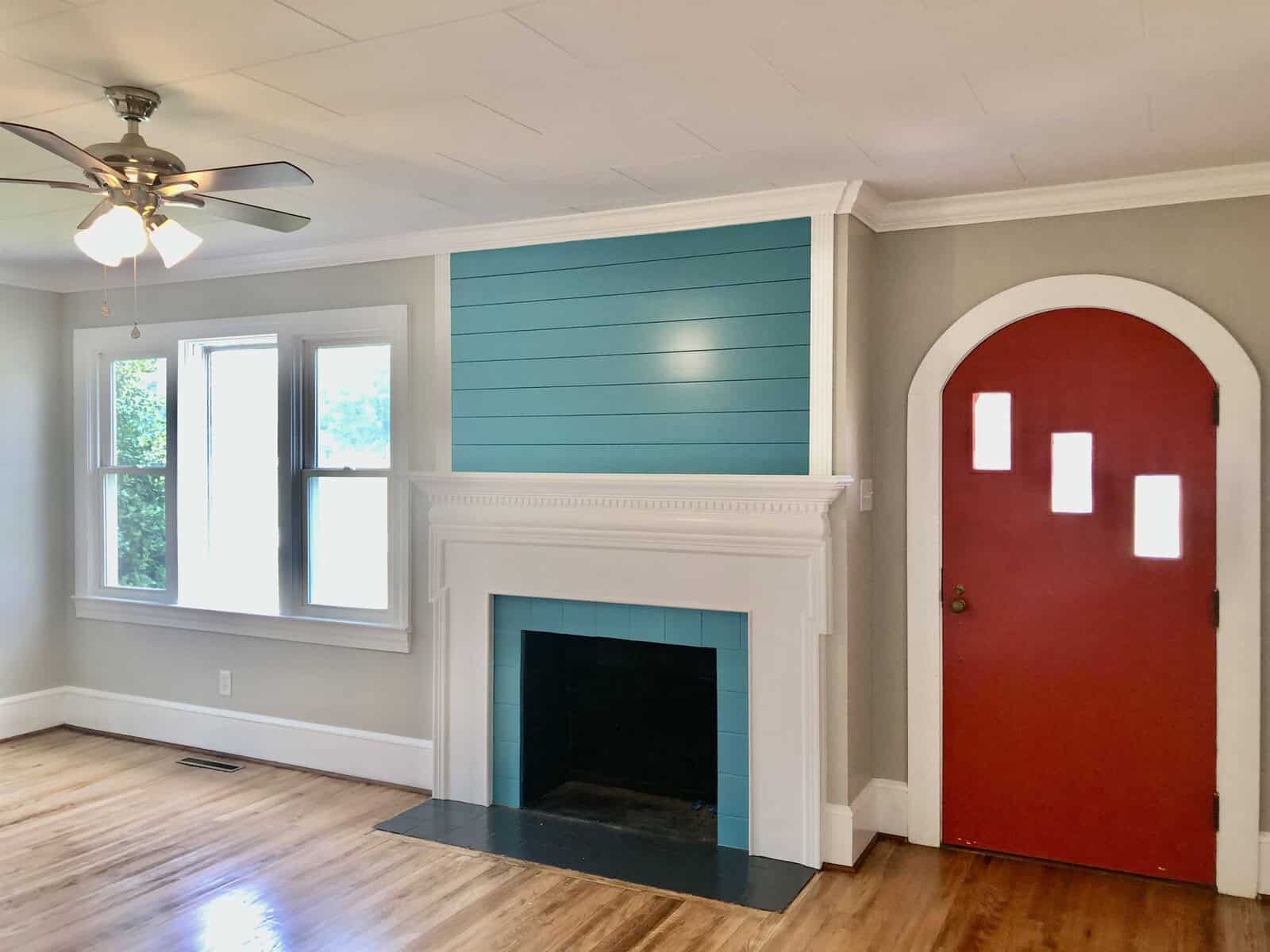 A freshly painted room featuring a white fireplace with a teal-colored accent wall above it, two windows to the left providing natural light, and a red arched door to the right.