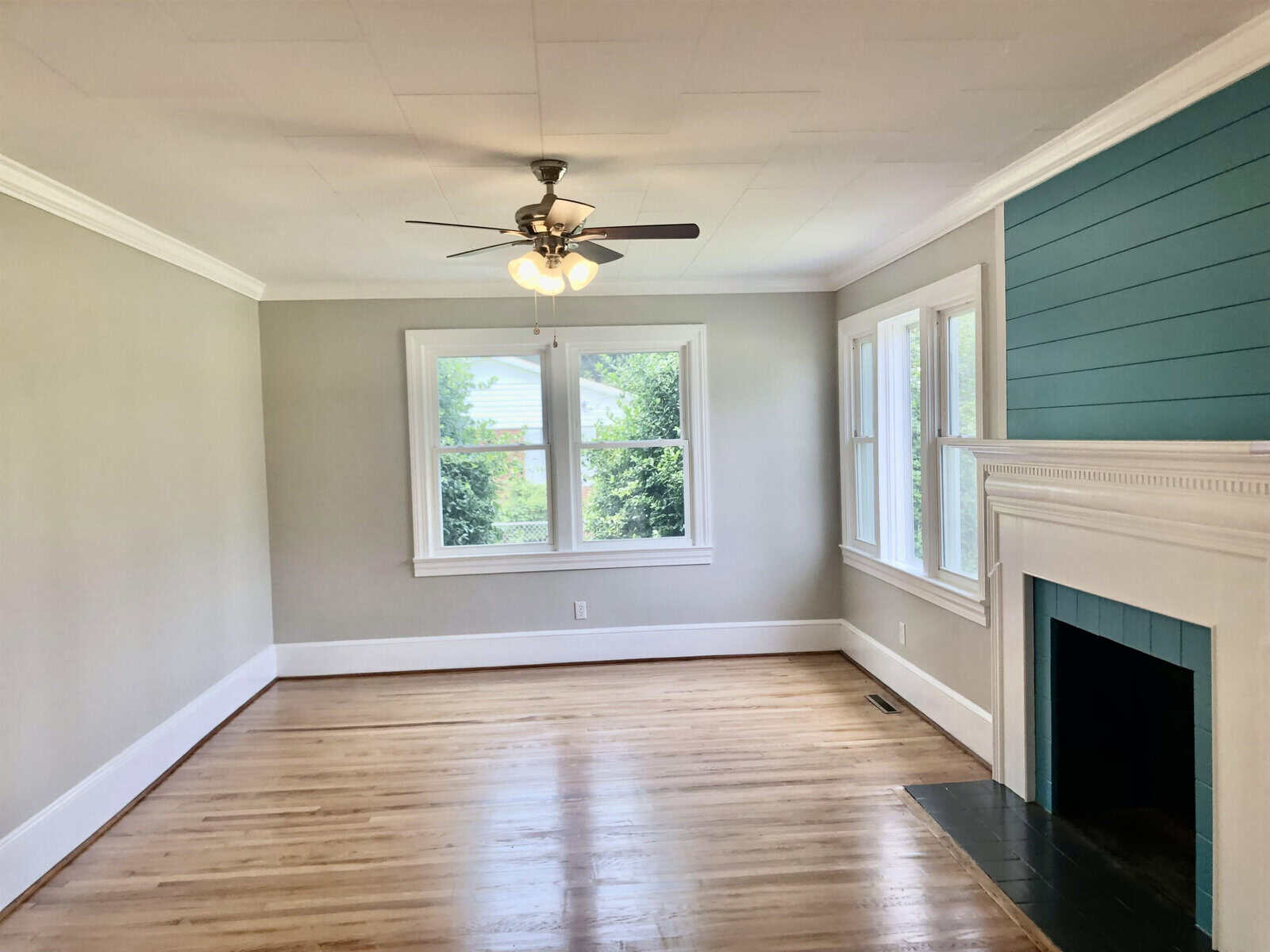 An empty room with hardwood floors, light gray walls, a white ceiling with a ceiling fan, a fireplace, and windows providing natural light.