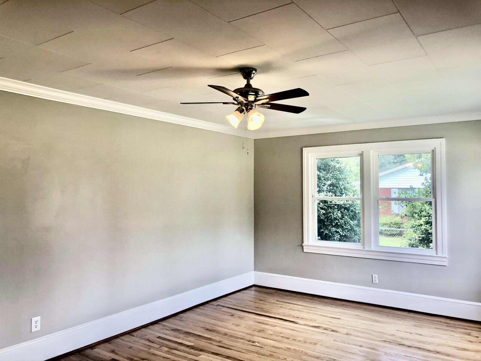 An empty room with gray walls, wooden flooring, a ceiling fan with lights, and a window showing an outside view.