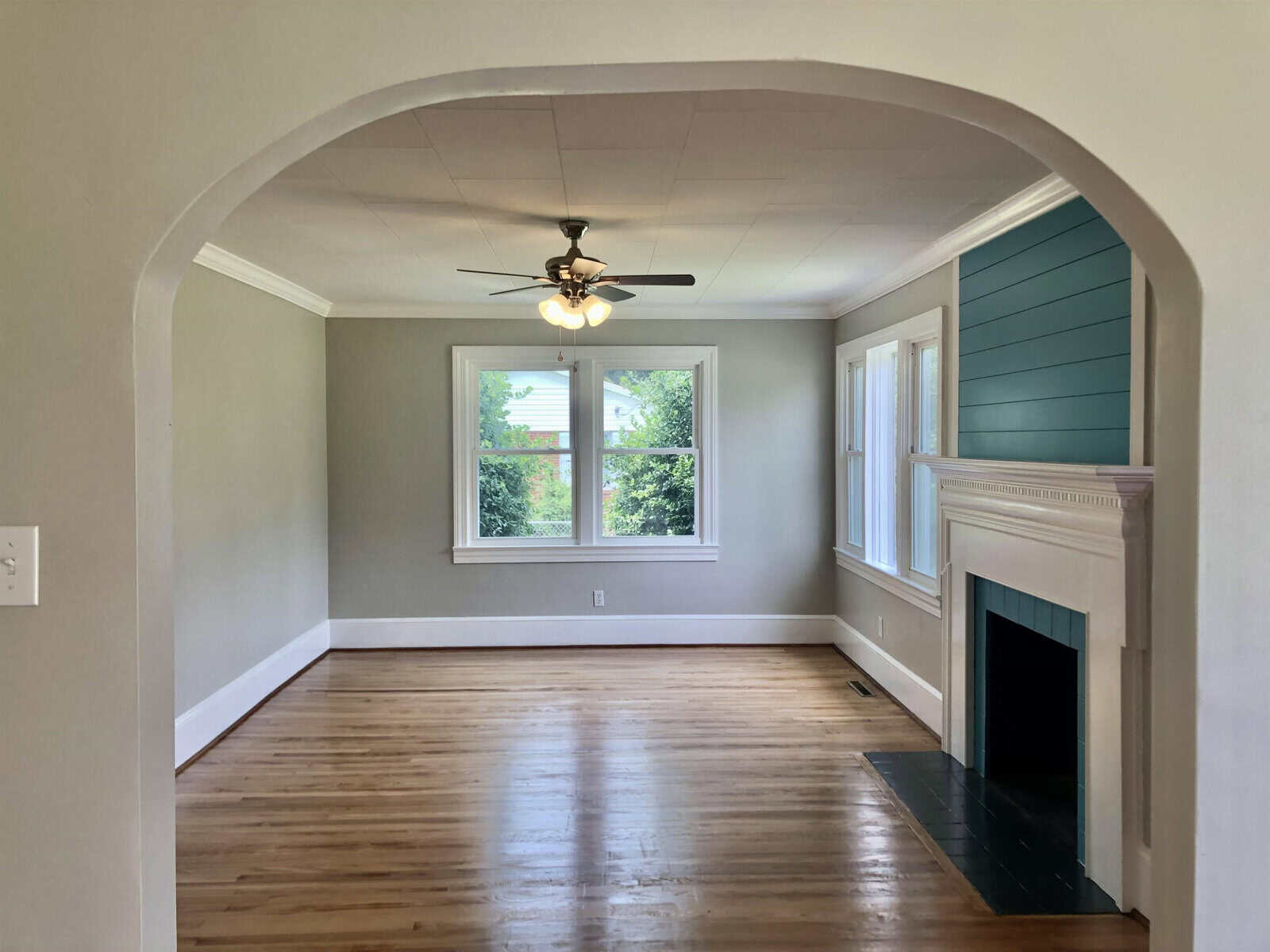 A spacious, empty room featuring hardwood floors, a white fireplace, and an archway entrance with a ceiling fan and natural light coming from the windows.