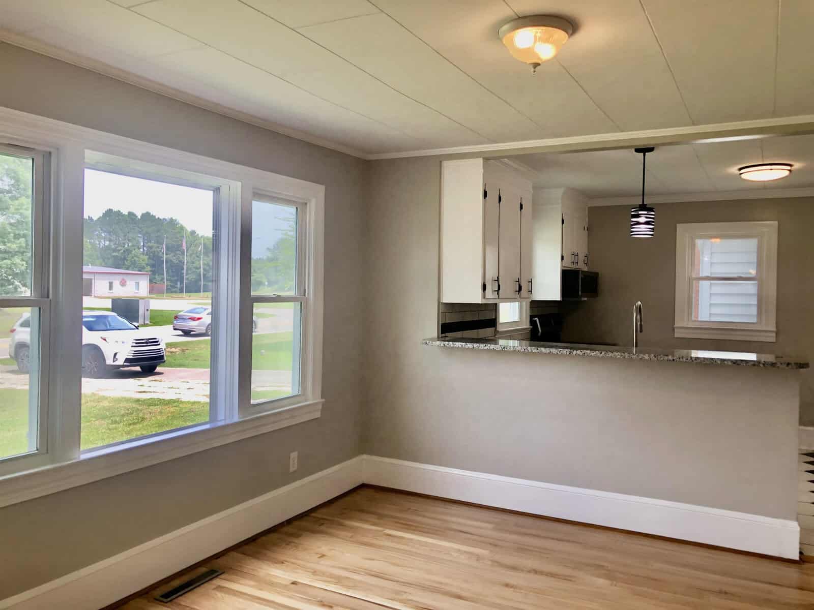 An empty room with hardwood floors, white walls, and multiple windows offering a view of the outside. the room extends into an open kitchen area with white cabinets and dark countertops, with a pendant light hanging overhead. a staircase is partially visible on the right, and a vehicle is parked outside one of the windows.