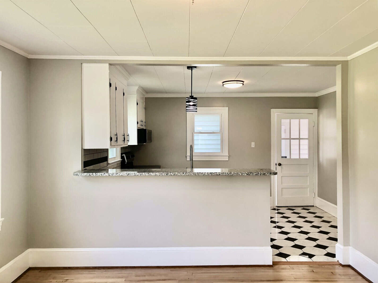 A modern kitchen interior featuring white cabinetry and a breakfast bar, with a pendant light above and black-and-white checkered flooring leading to an adjoining room with a white door.
