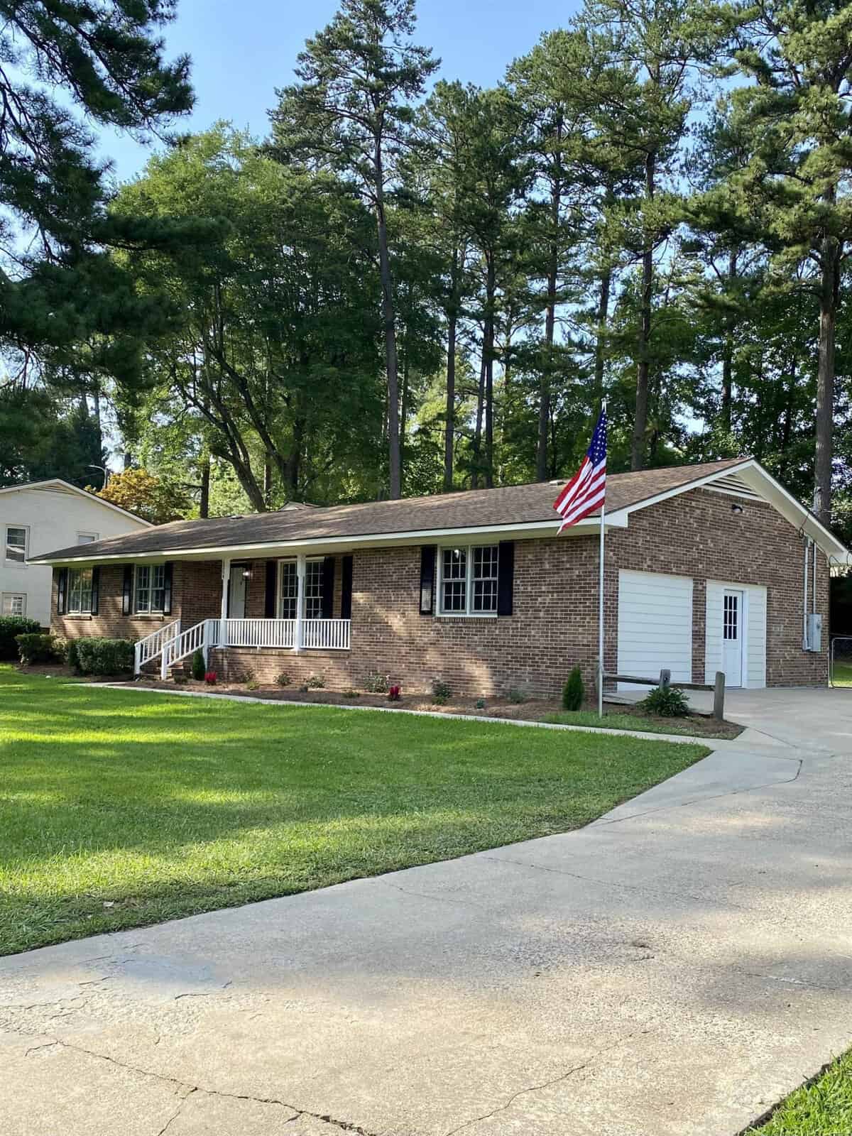 A single-story brick house with a white door and garage, featuring a neatly manicured lawn and an american flag displayed on a flagpole.