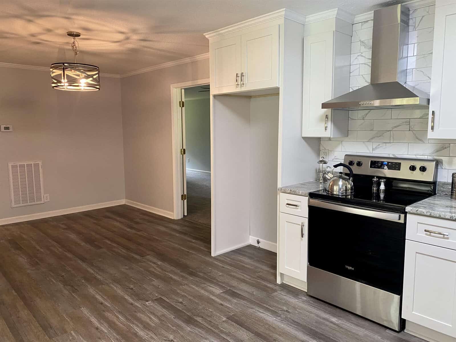 A modern kitchen with white cabinets, stainless steel appliances, and gray wood-patterned flooring, leading to an adjacent room through an open doorway.