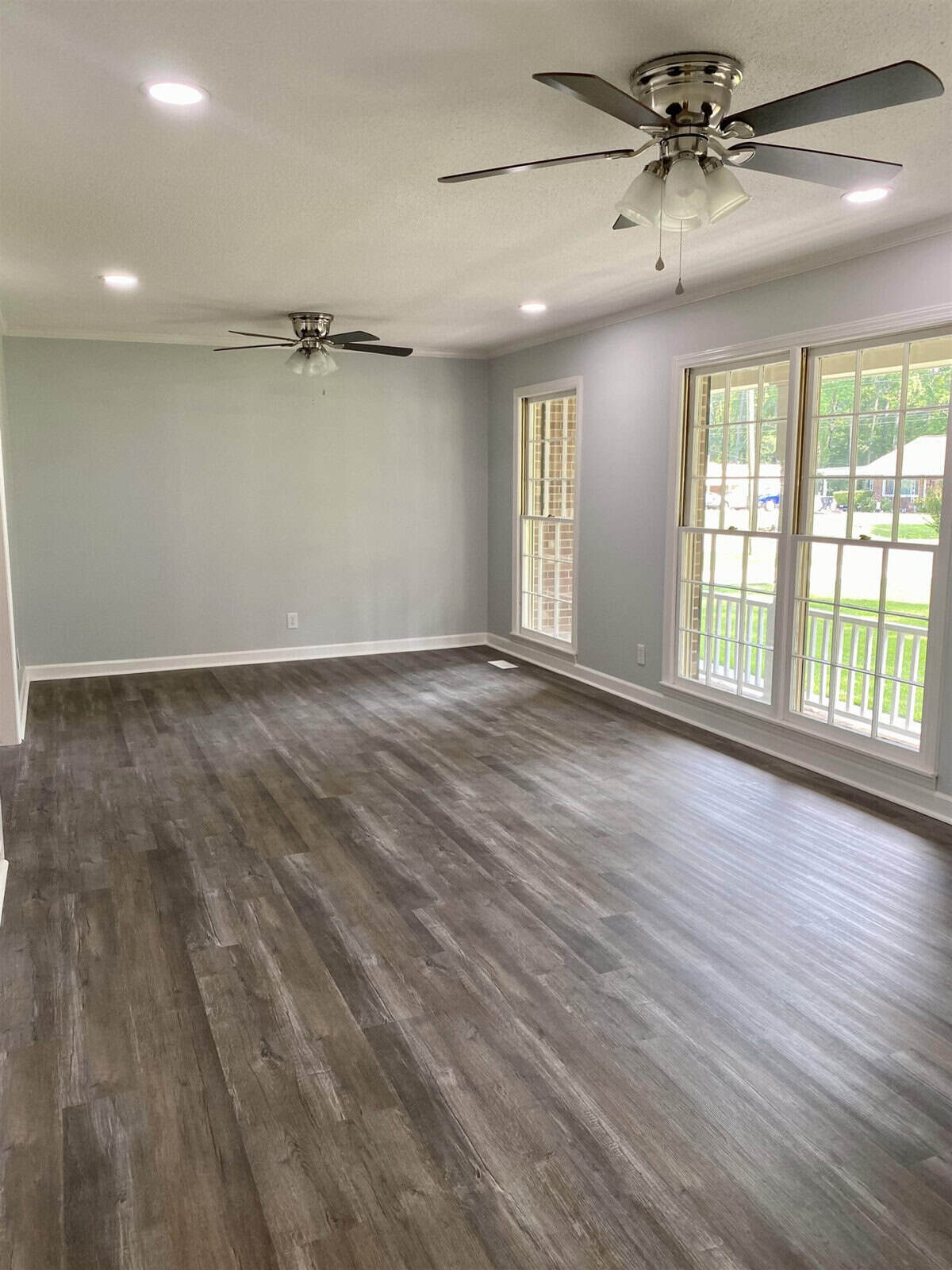 An empty room with gray walls, wooden flooring, two ceiling fans, and natural light coming in through the windows.