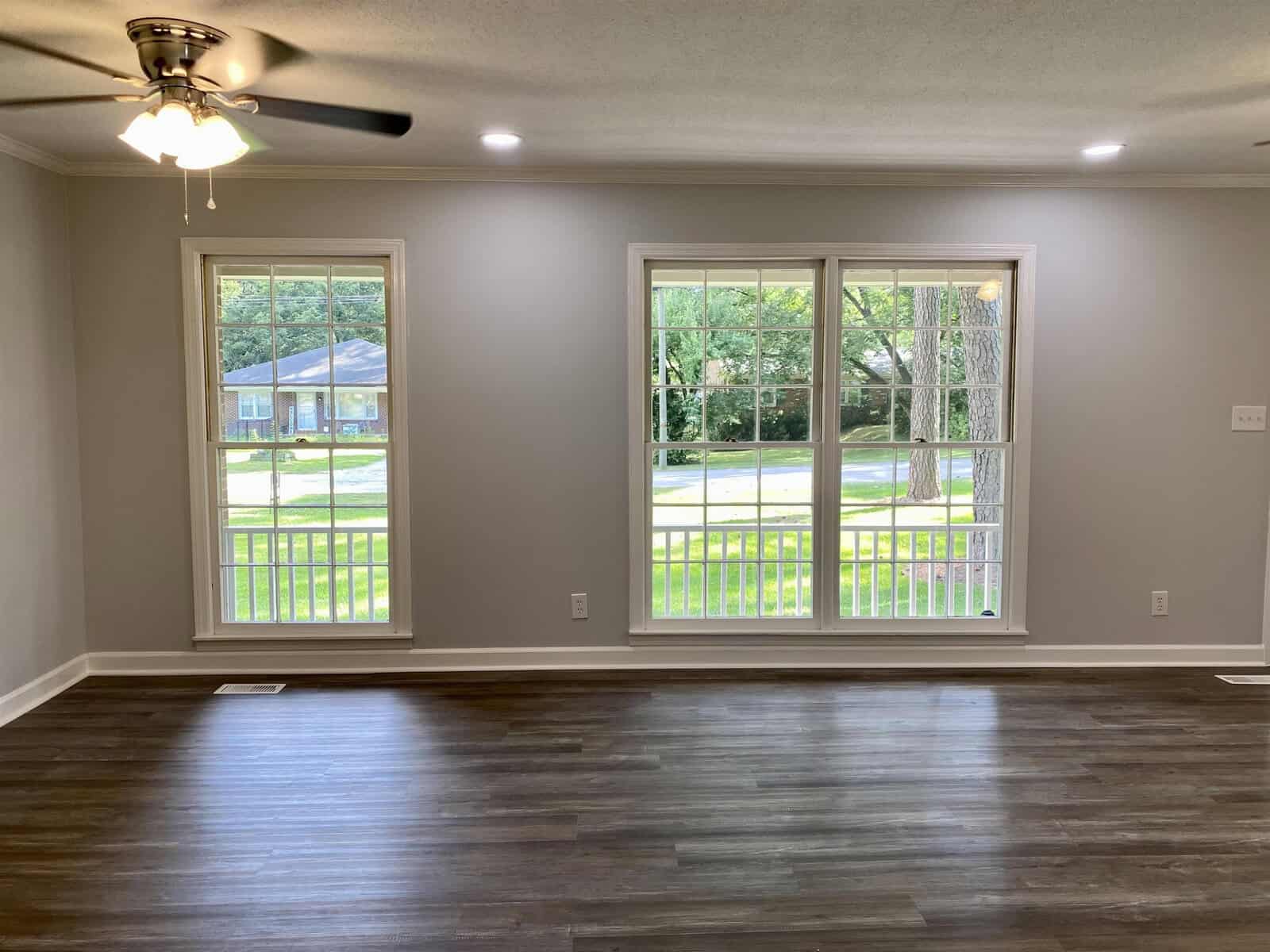 An empty room with gray walls and dark hardwood flooring featuring a ceiling fan and three large windows with a view of the street outside.