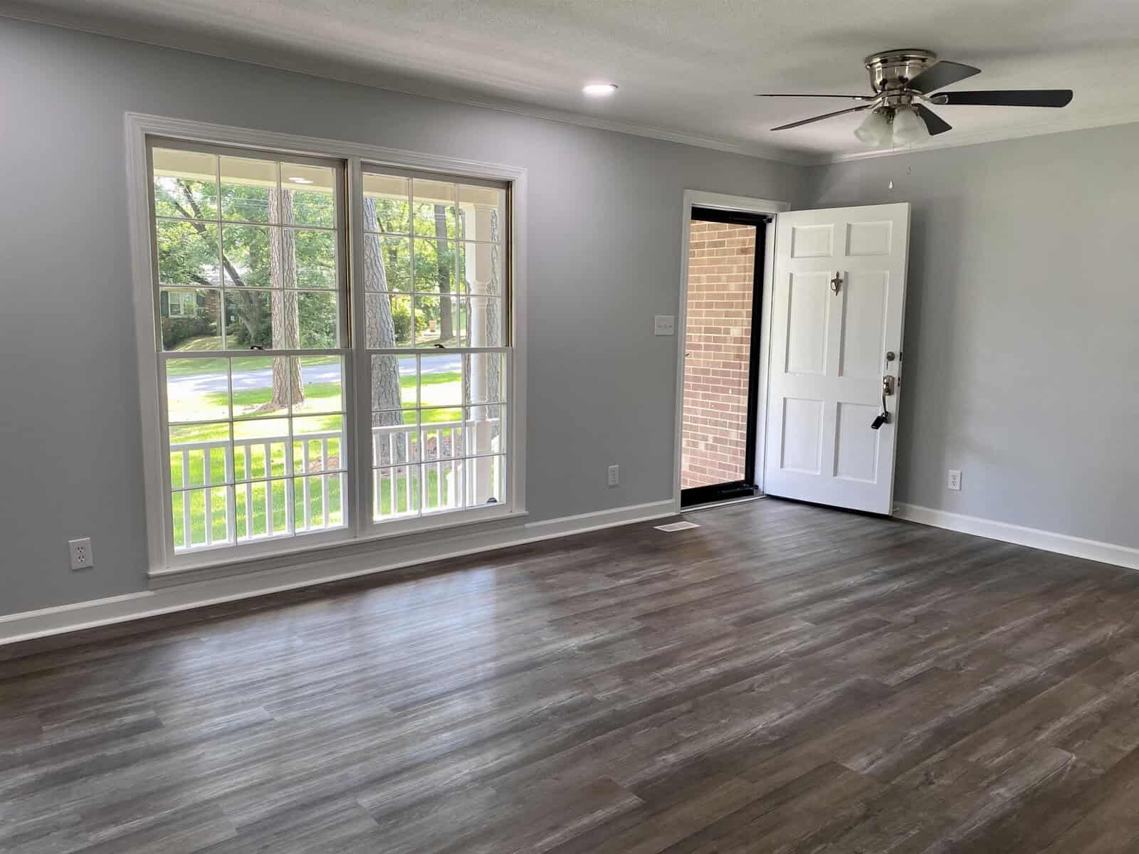 A spacious, empty room with grey flooring, a large window, white walls, a ceiling fan, and an entrance door.