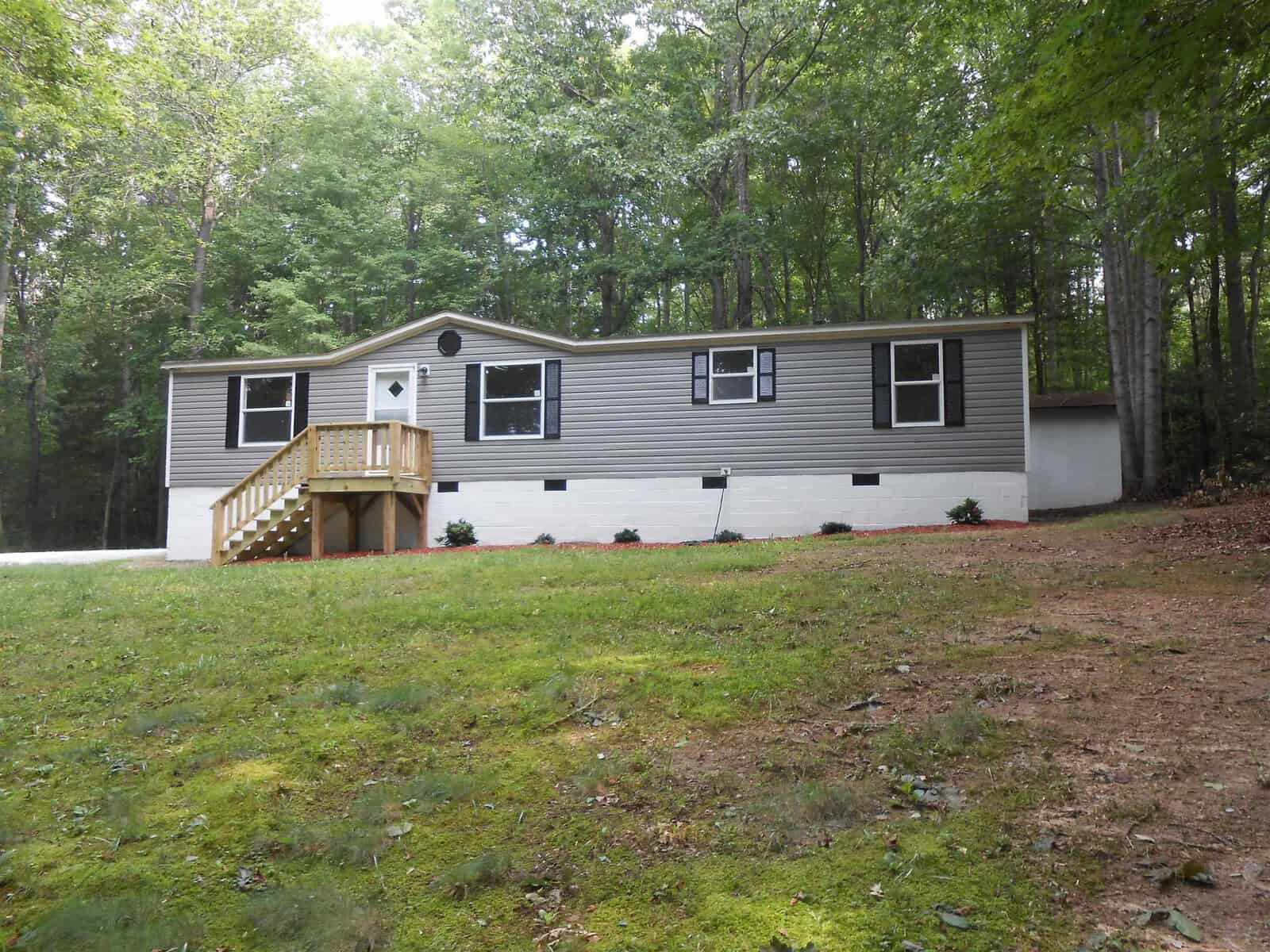 A single-story manufactured home with a gray exterior and white trim, featuring a small wooden staircase and deck at the entrance, set on a cleared lot surrounded by trees.