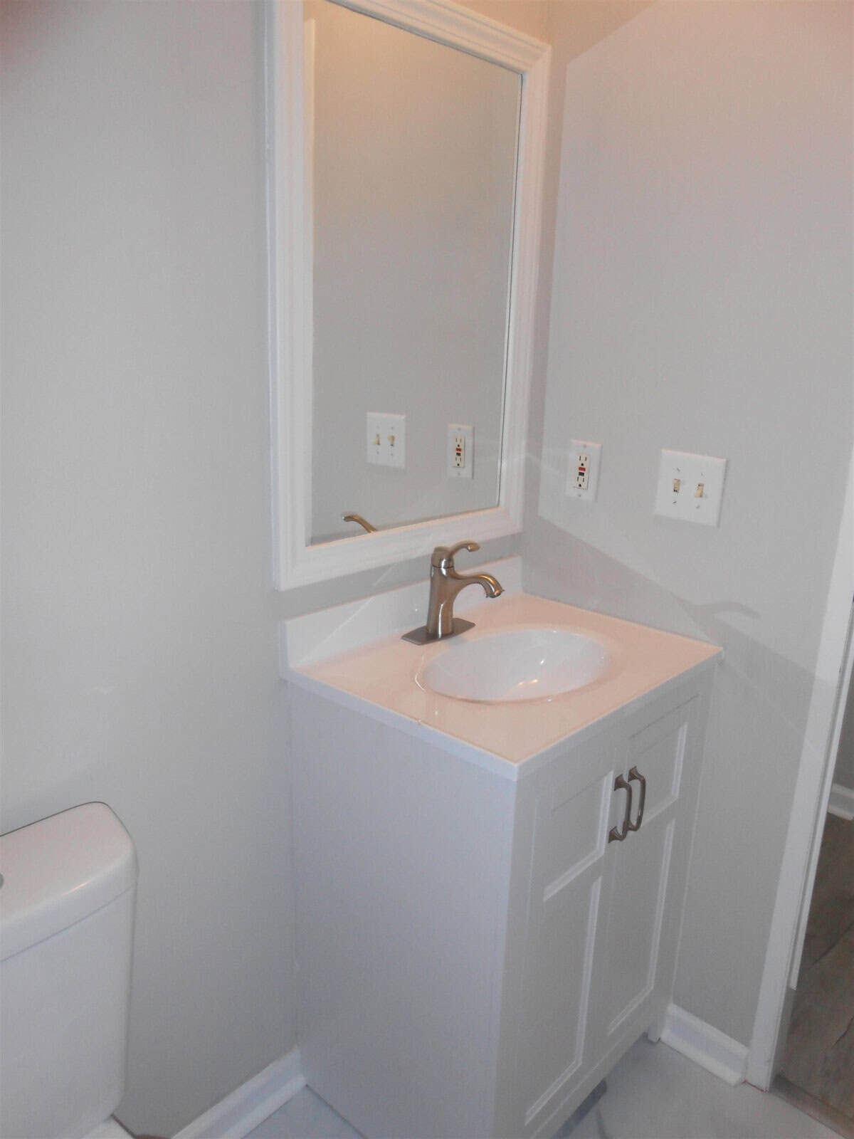 A small bathroom with white walls featuring a single vanity with a sink and mirror, next to a white toilet, with visible electrical outlets on the wall.