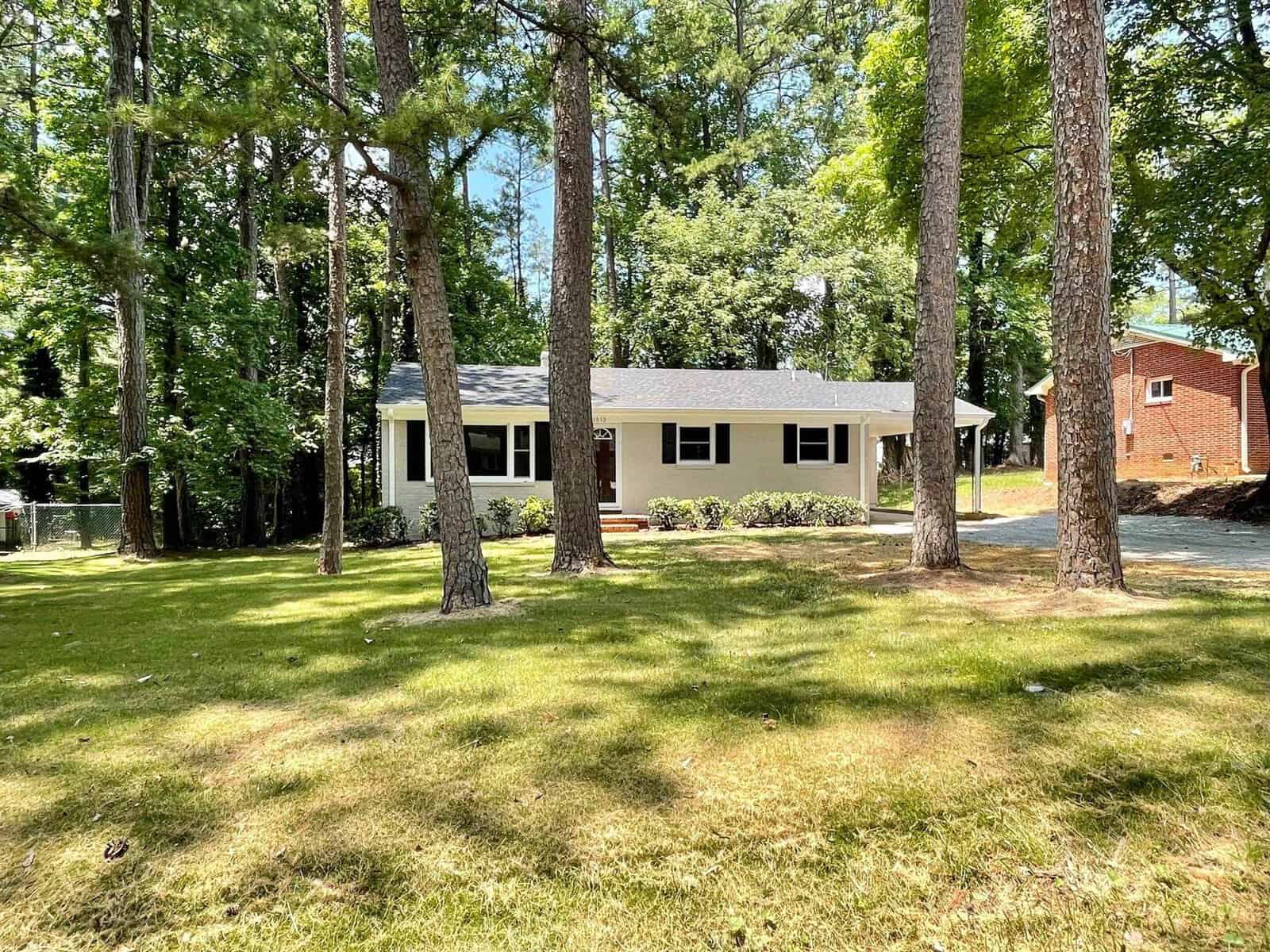 A single-story white house with a dark roof surrounded by tall pine trees on a sunny day with a well-manicured lawn in the foreground.