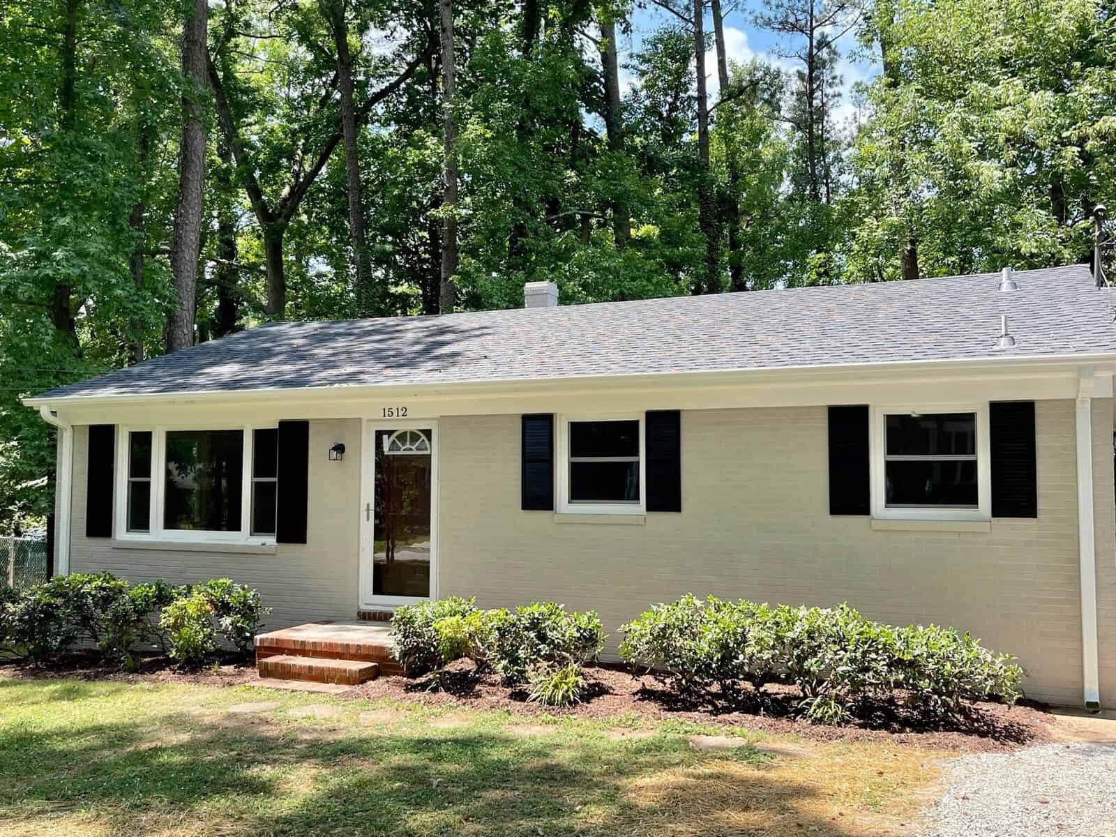 A single-story house with a beige exterior, black shutters, and a small front porch, surrounded by green trees and shrubs.