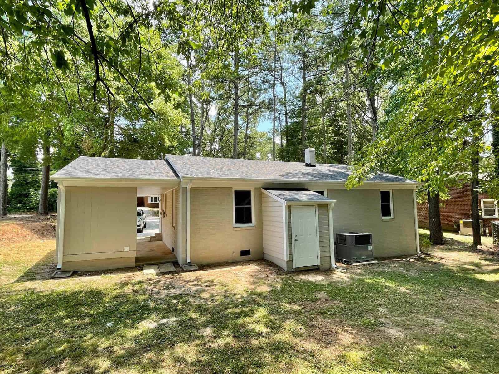A single-story house with beige siding and a brown roof, featuring an external hvac unit and surrounded by tall trees.