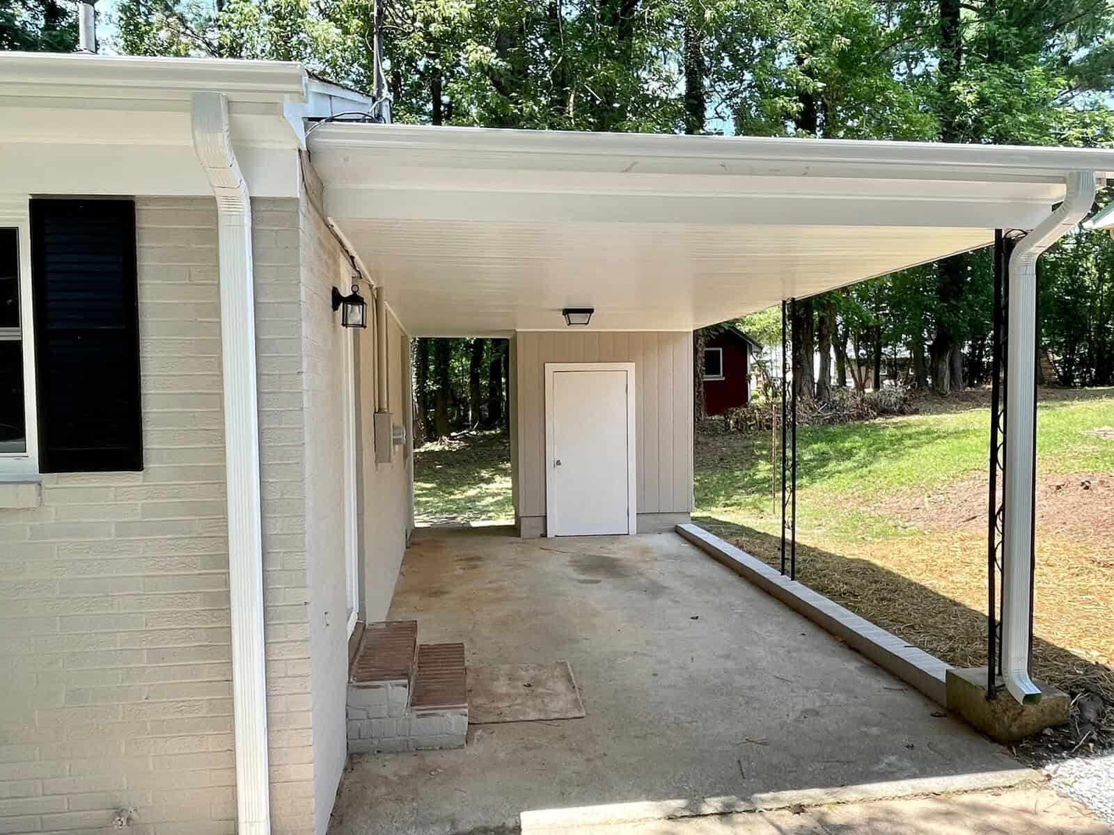 A white carport attached to a brick house, covering a concrete slab with a single door leading into the home.