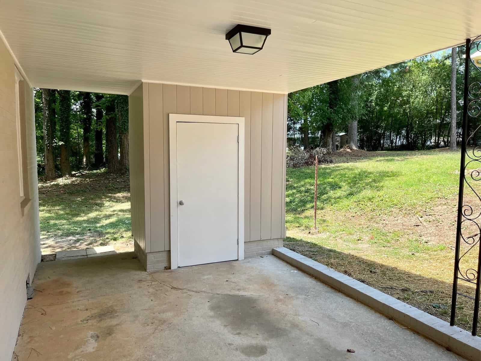 A white door at the end of a covered concrete patio area with a view of trees and grass in the background.