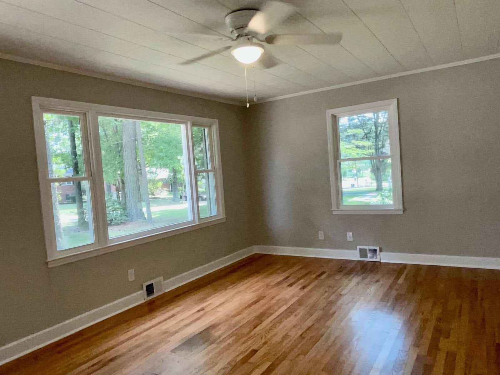 An empty room with polished wooden floors, white walls, a ceiling fan, and three large windows providing a view of trees outside.