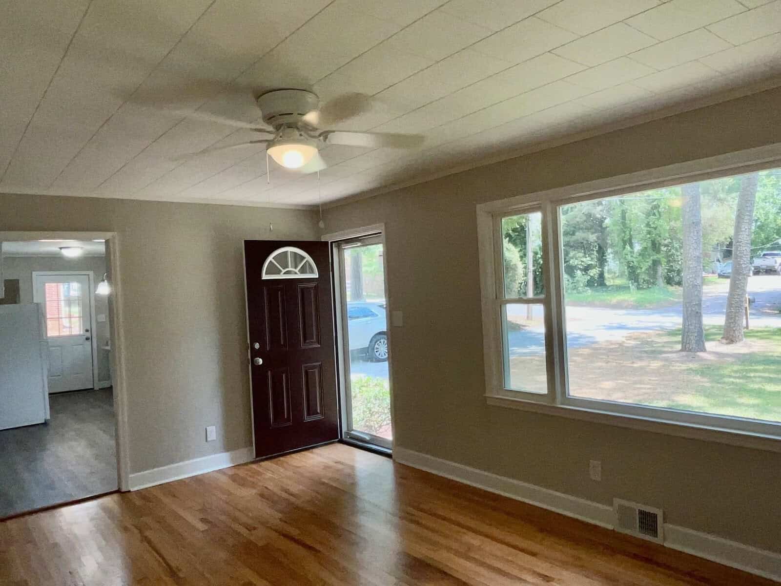 An empty room with hardwood floors, a white ceiling with visible patterns, a ceiling fan, and a brown door with an arched window at the top. there is a large window on the right wall providing a view of the outside area and a parked car.