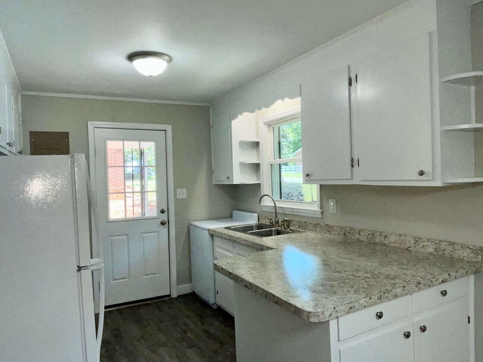A small, modern kitchen with white cabinetry, granite countertops, and stainless steel appliances, viewed from an angle that includes the entrance door with a view of the outside area.