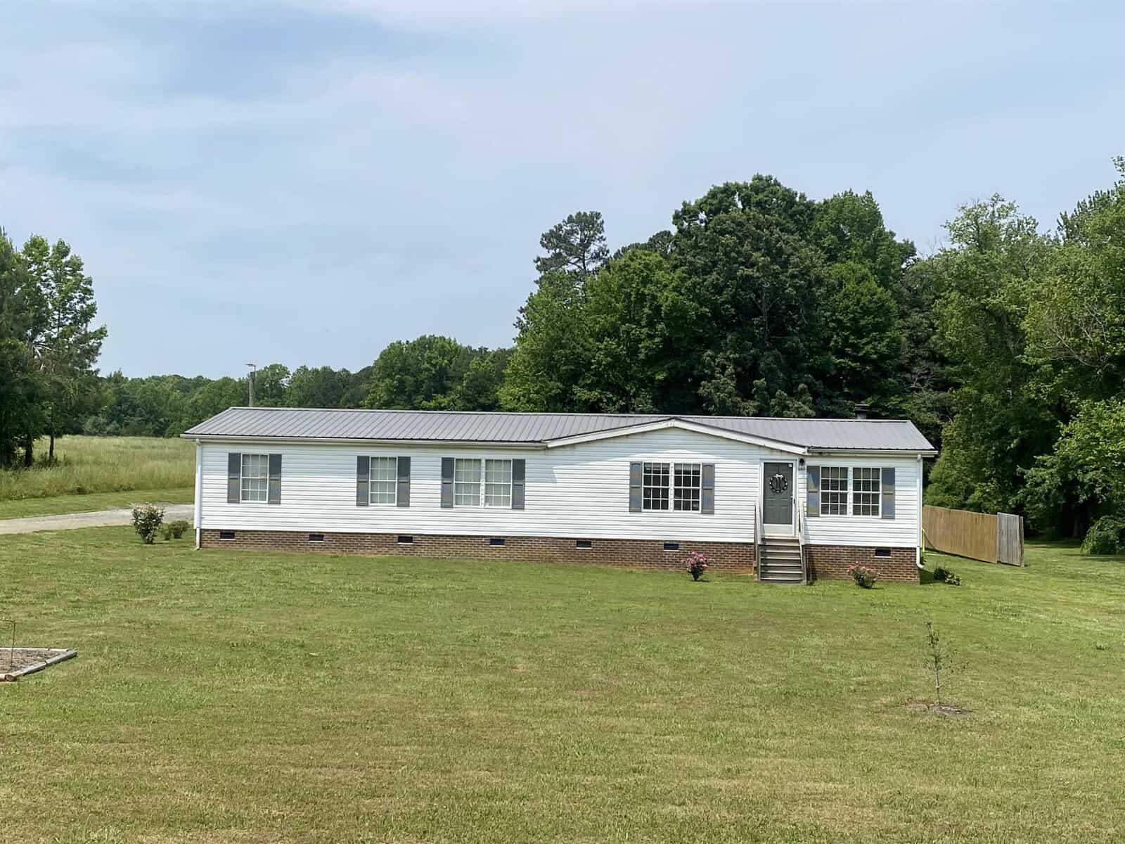 A single-story white house with a gray metal roof and brick foundation, located at 3627 & 362 Cannady Mill Road in Kittrell, is surrounded by a large, neatly mowed lawn. The house has several windows and a front door with steps leading up to it. Trees and foliage are visible in the background.