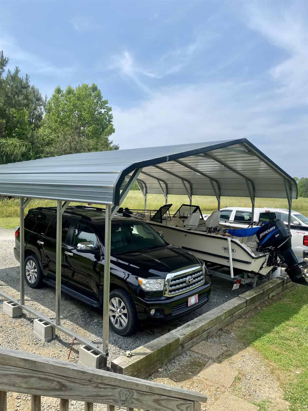 A black SUV and a white motorboat are parked under a metal carport on a gravel driveway. The carport is supported by cinder blocks. A white pickup truck is partially visible in the background. The area is surrounded by greenery and trees under a clear sky.