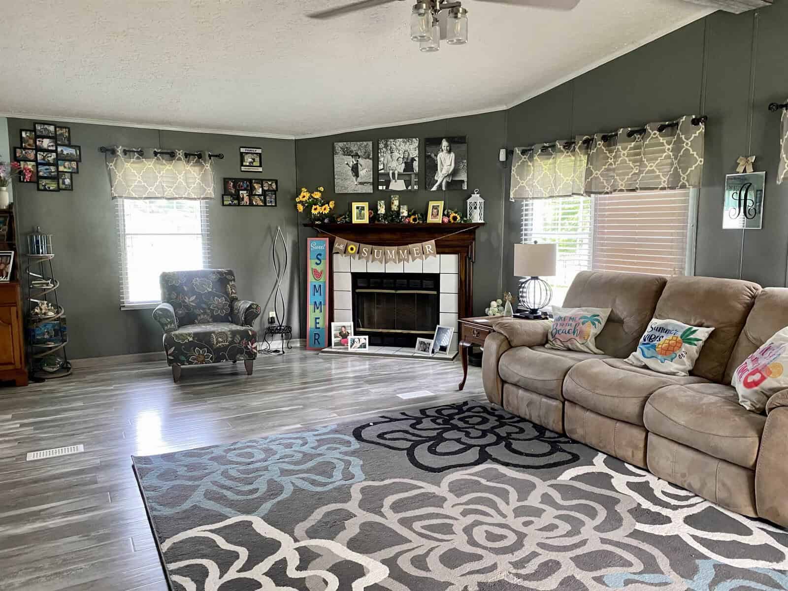 A cozy living room with gray walls features a beige sectional sofa with floral pillows, a fireplace decorated with "SUMMER" letters and family photos, an armchair next to a window, patterned curtains, and a gray floral area rug on wood flooring.